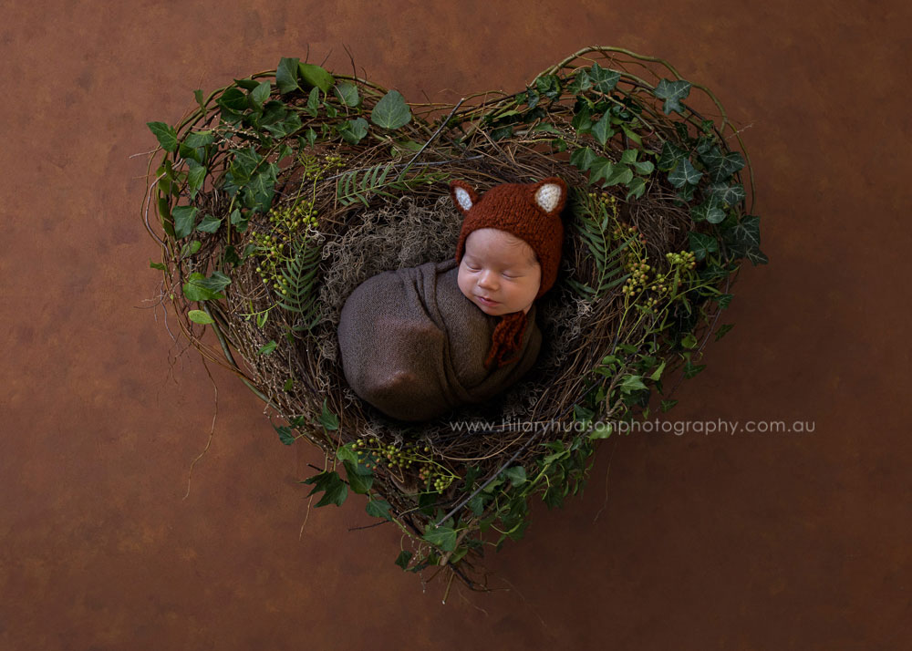 Cute baby with a fox beanie, snoozing on a vine heart nest with greenery and leaves