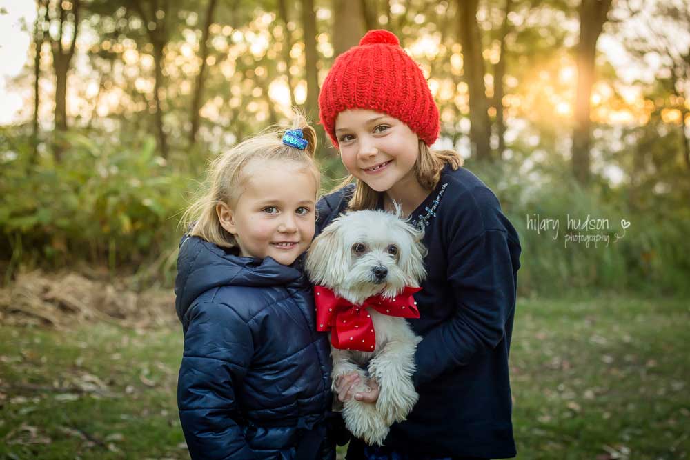 Two sisters cuddling their adorable little white dog who is wearing a red ribbon.