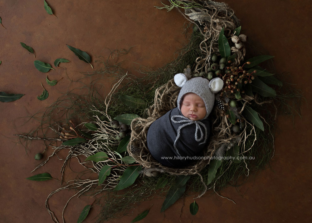 Baby in a steel blue wrap and bear beanie, sleeping in a nest of nature elements and leaves