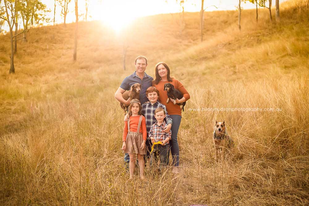 Family of 5, with 3 dogs, standing in a rural field lit with golden afternoon light