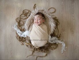 smiling baby wearing a beige/neutral wrap, lying in a wicker basket with jute and lace